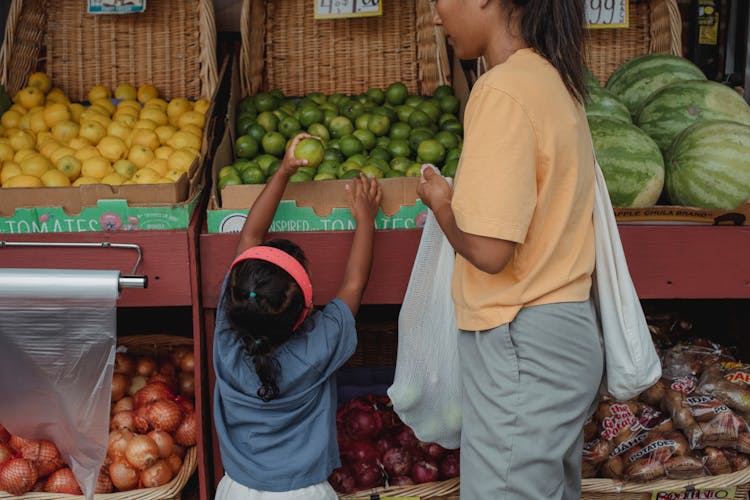 Ethnic Girl Choosing Fruit From Box In Bazaar With Mother