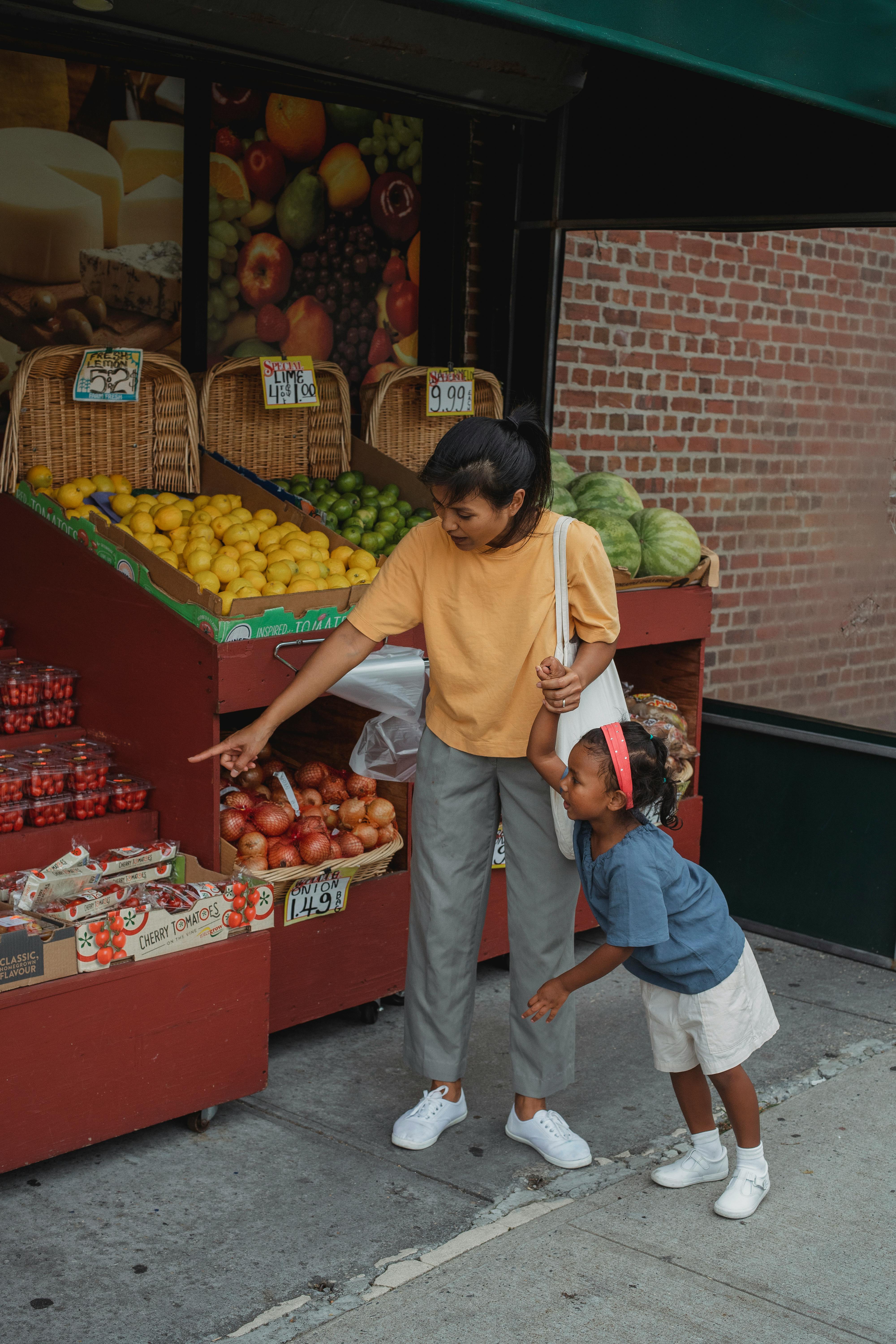 ethnic woman pointing at vegetables in market with daughter