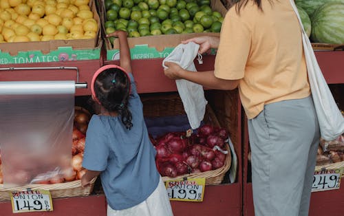 Back view of crop anonymous mother with reusable bag and girl choosing limes from box