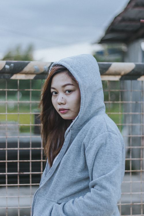 Free Young woman in gray hoodie standing by a fence outdoors in the Philippines. Stock Photo