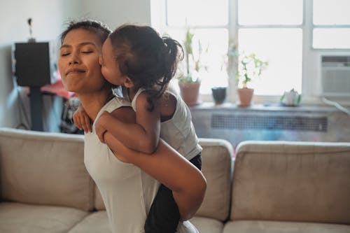 Free Side view positive young Asian woman piggybacking adorable daughter and getting cheek kiss while having fun together in light living room Stock Photo