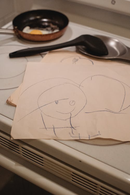 Kids drawing placed on stove near frying pan with egg