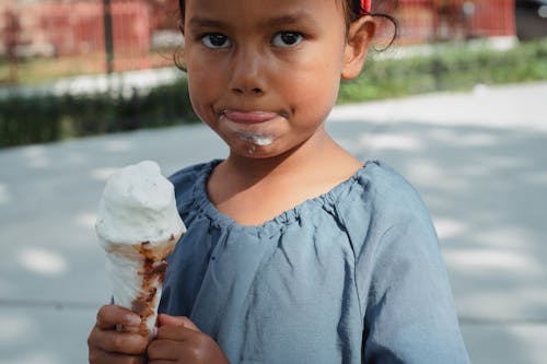 Crop adorable little Asian girl in casual clothes eating ice cream and looking at camera