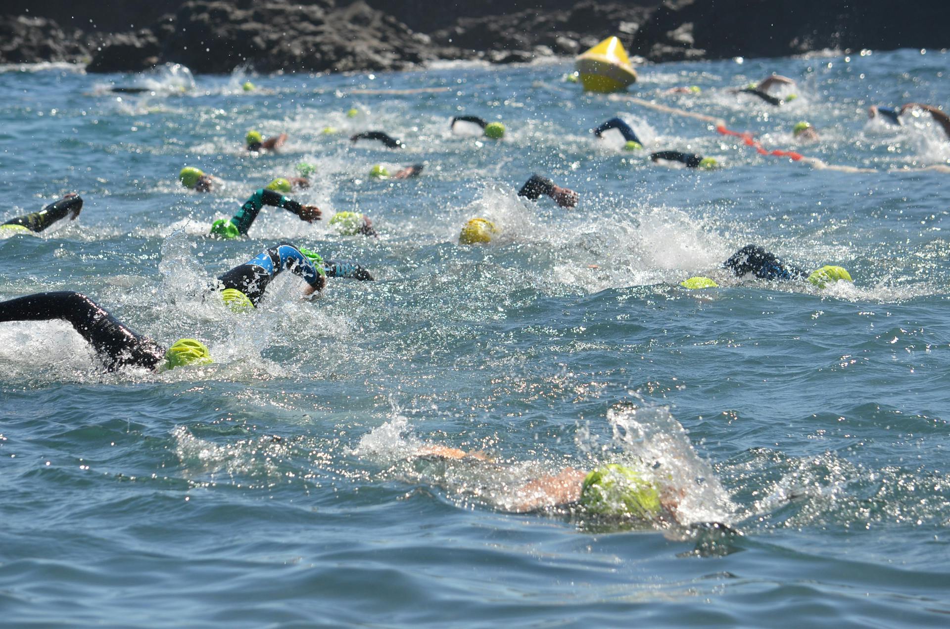 Competitive swimmers in a triathlon swim race, splashing through ocean waters, wearing swim caps.