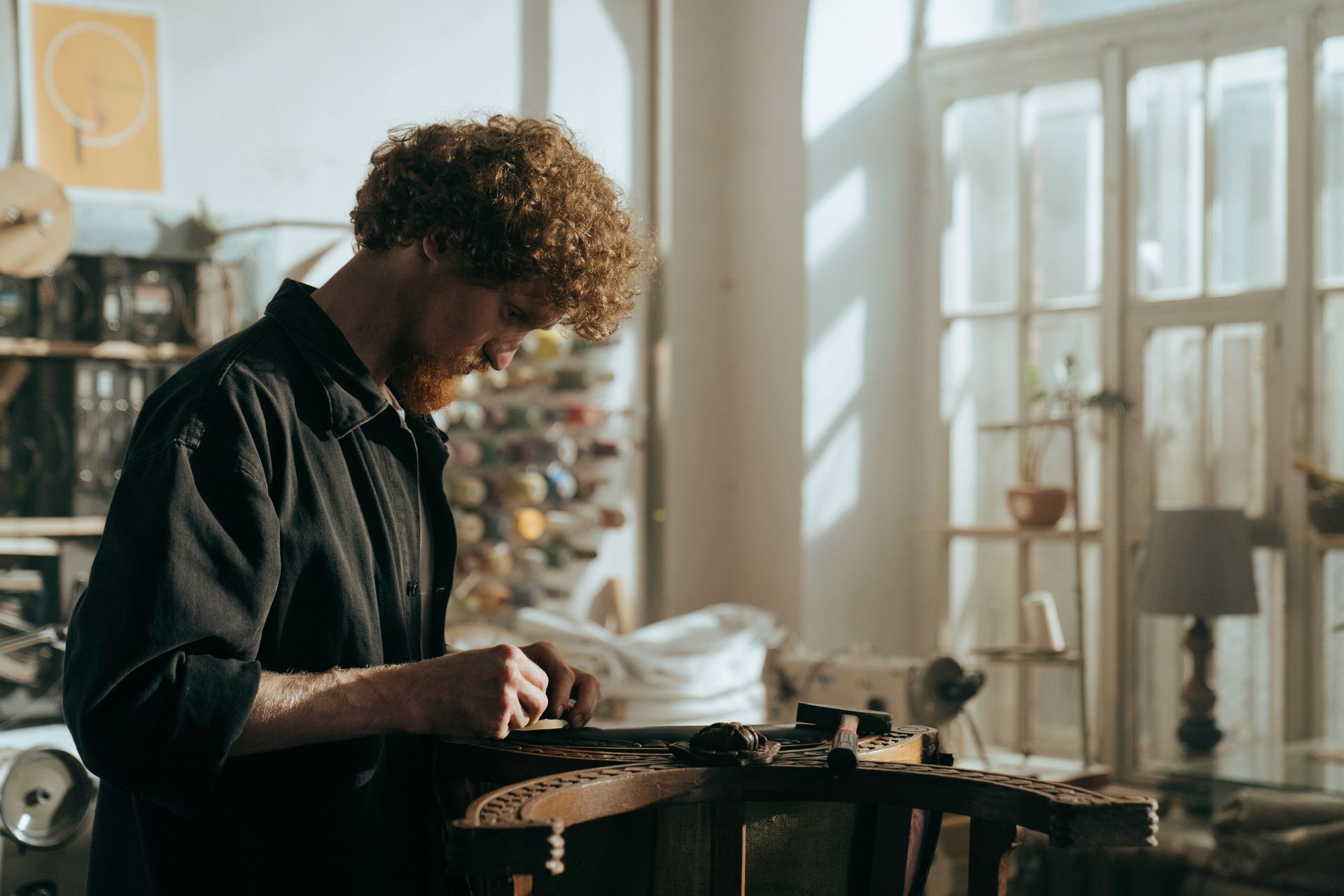 A redheaded craftsman focuses on restoration work in a sunlit workshop, surrounded by tools and materials.