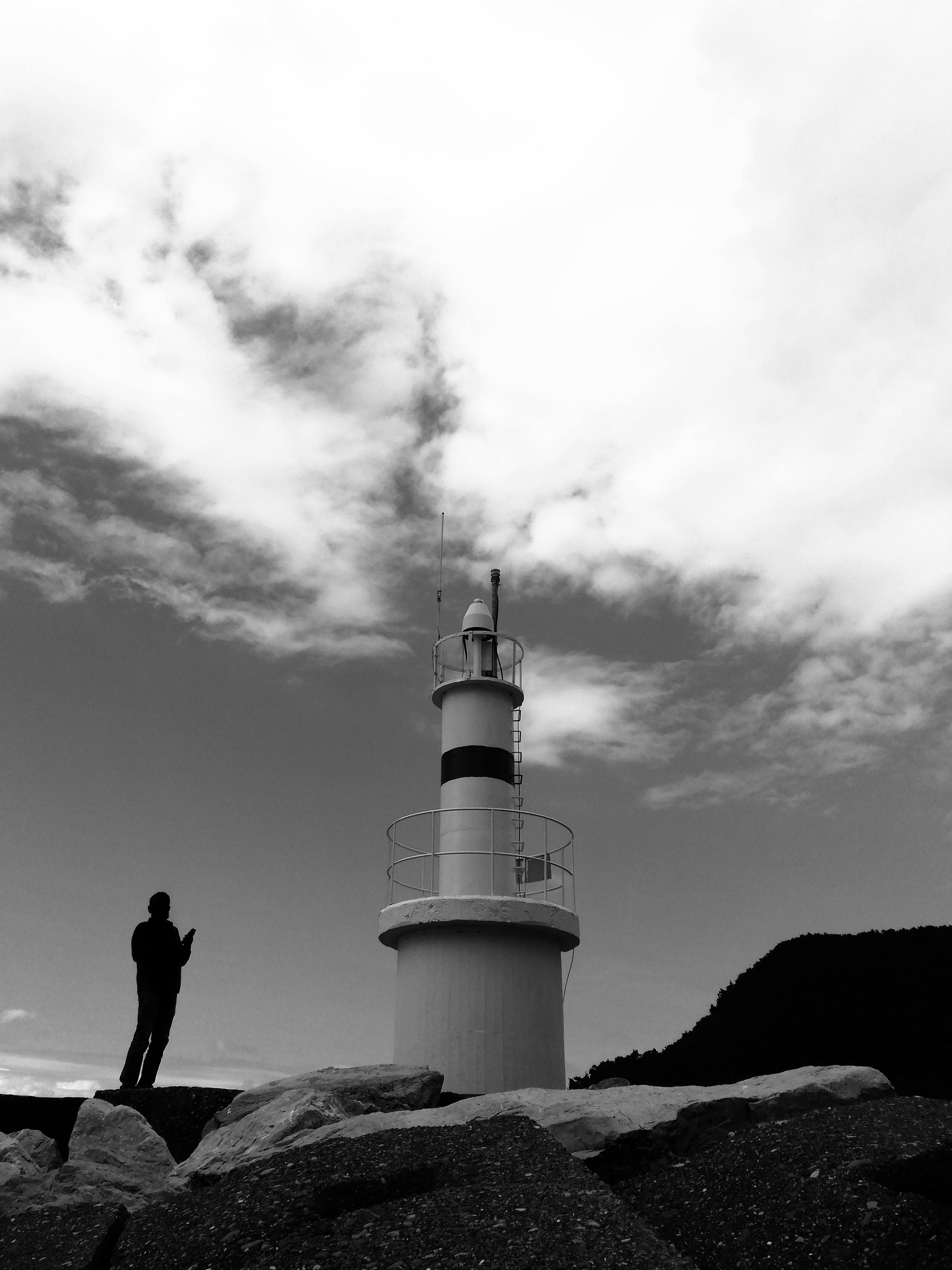 A silhouetted man stands near a towering lighthouse in black and white, under a dramatic cloudy sky.