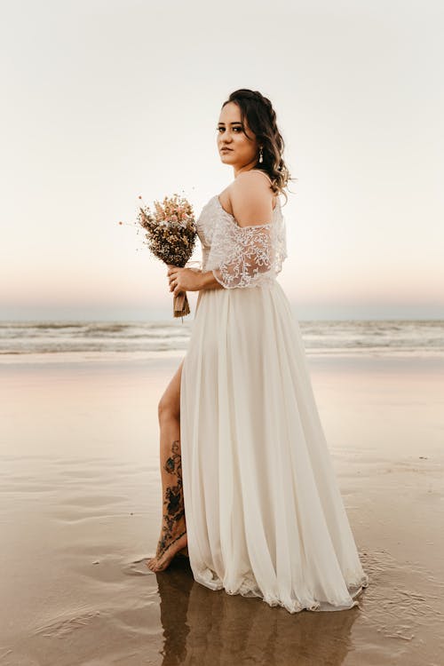 Barefoot tattooed bride in white dress with open shoulders standing on sandy shore with flower bouquet in hand