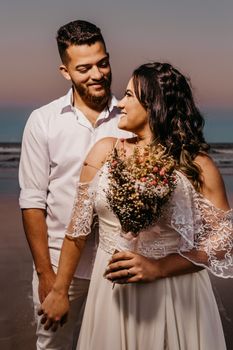 Content groom and bride in white wedding outfits standing on sandy beach holding hands and looking at each other