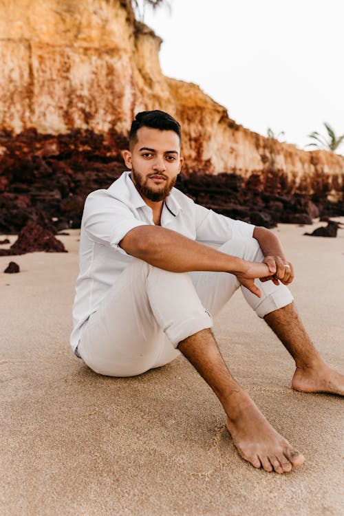 Handsome young ethnic male wearing stylish white clothes sitting on sandy shore and embracing knees while looking at camera