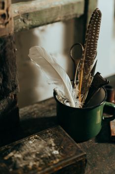 木桌上放着一个复古马克杯，里面装满了羽毛和旧工具，场景质朴