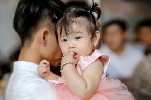 Free Adorable Asian toddler with ponytails in pink dress sitting on hands and sucking finger Stock Photo