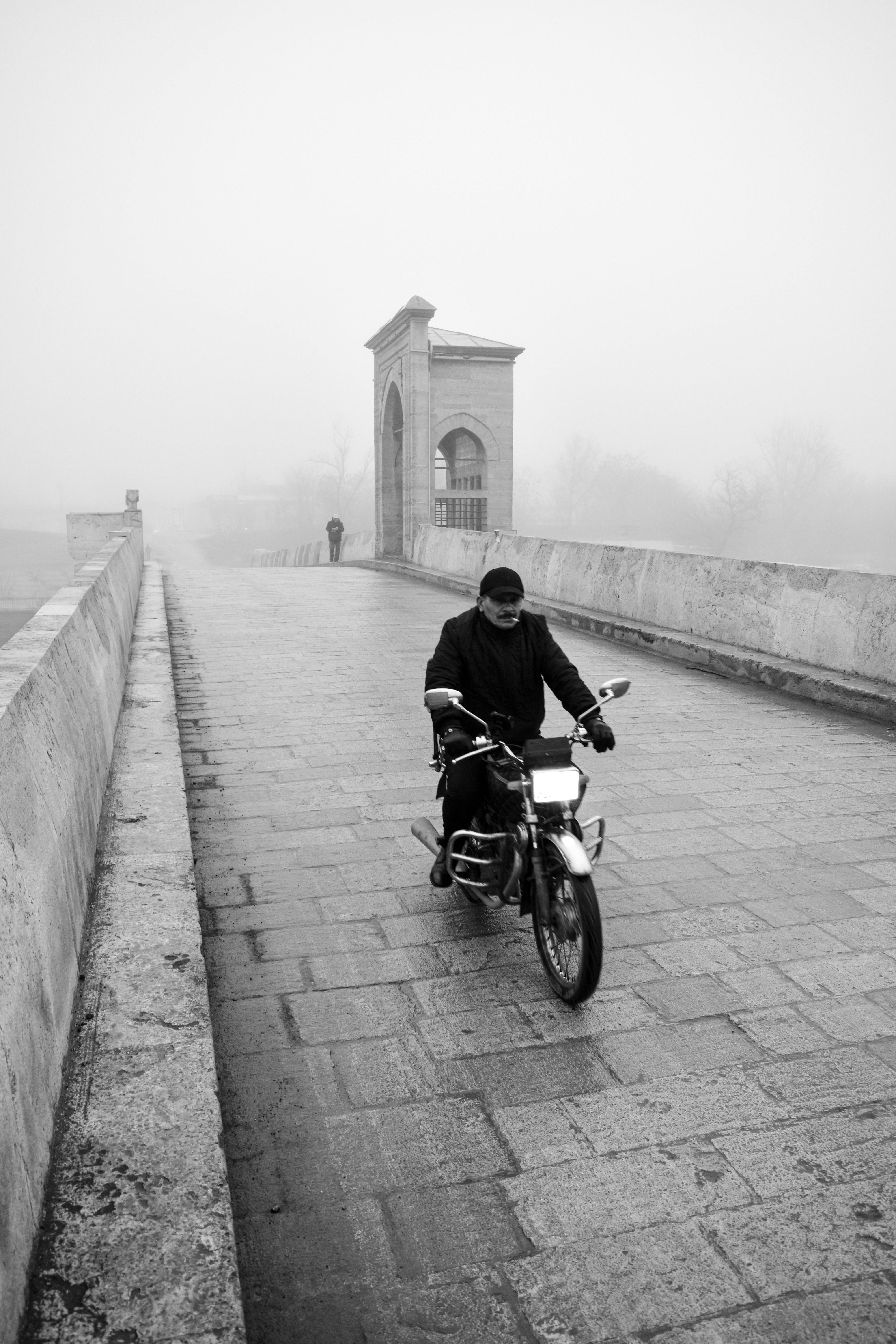 Black and white photo of a man riding a motorcycle on a foggy bridge in Eskişehir, Turkey.