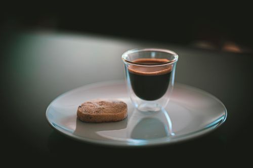 Free Close-up of a double-walled shot glass with espresso and a round biscuit on a glossy white plate. Stock Photo