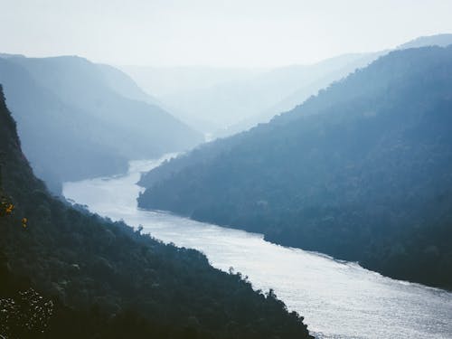 Scenic view of rippled river between ridges with trees under light sky on misty day