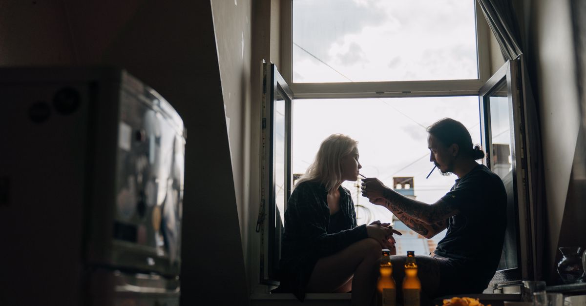 A couple enjoying a quiet moment on a windowsill with beer bottles and a cigarette indoors.