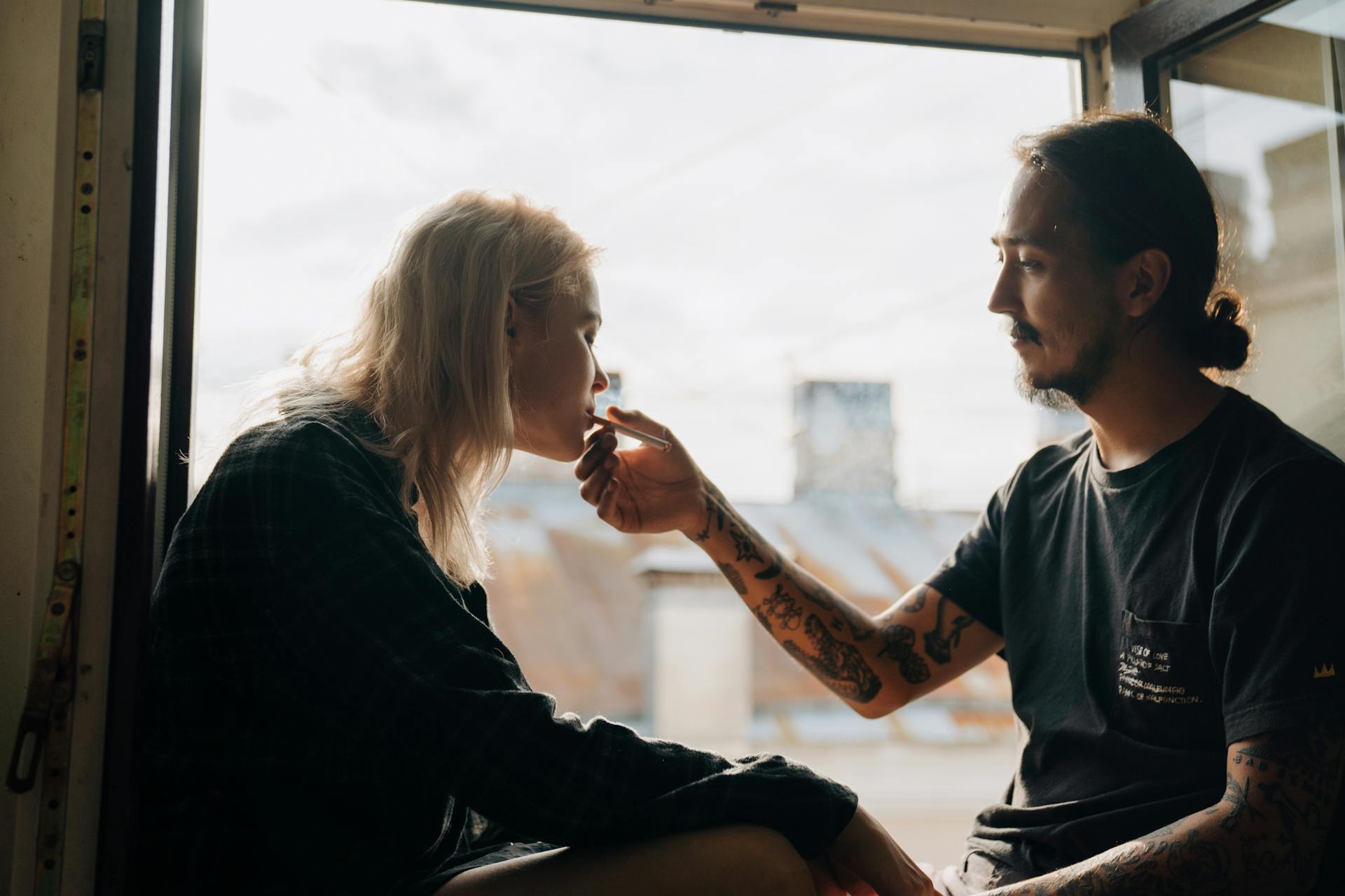 A young couple shares a moment by the window as they smoke together, enjoying a bonding time.