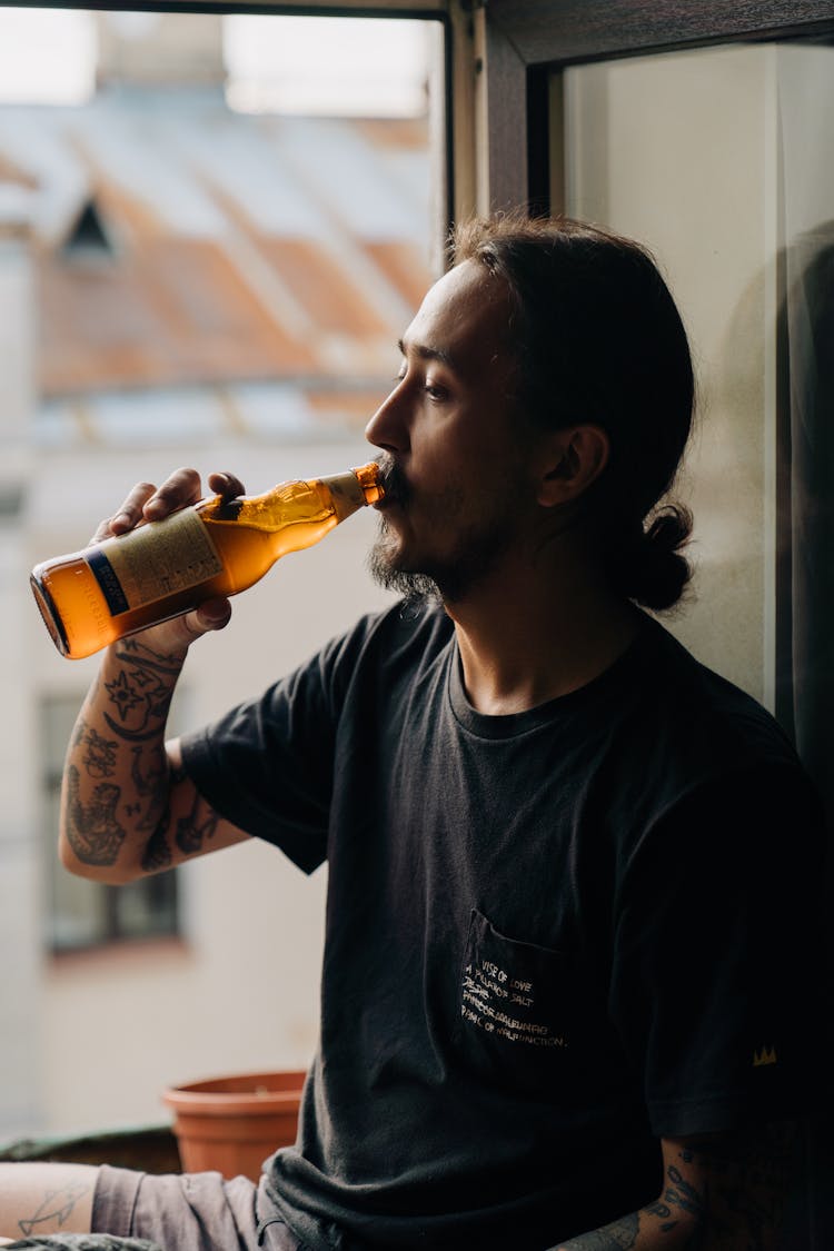 A Tattooed Man Drinking A Beer Bottle By A Window