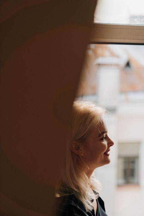 Free A woman with blonde hair sitting by a window, smiling and wearing a checkered shirt indoors. Stock Photo
