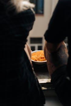 A bowl of potato chips on a table surrounded by people indoors, highlighting a casual snack time atmosphere.