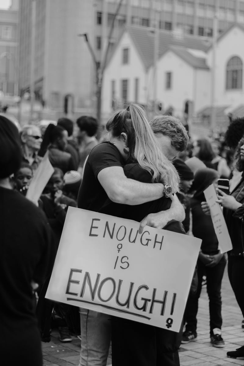 Black and white of unrecognizable couple hugging and standing on street with sign with ENOUGH IS ENOUGH inscription against crowd of people