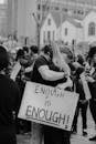 Black and white of unrecognizable couple hugging and standing on street with sign with ENOUGH IS ENOUGH inscription against crowd of people