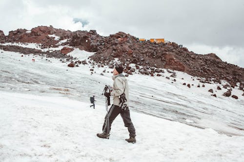 Free Full body of male traveler in warm outerwear walking with trekking sticks on snowy mountain ridge in daytime Stock Photo