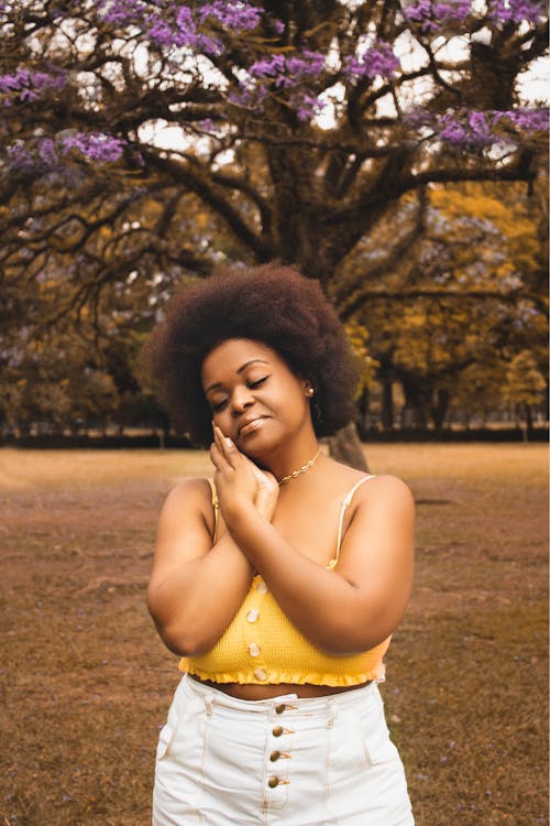 Plump African American female with afro hairstyle in casual clothes standing against tree with blooming violet flowers