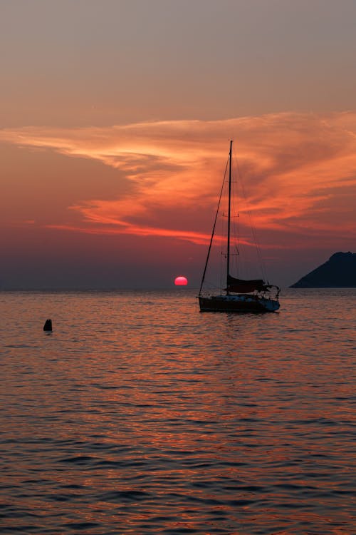 Scenery view of sailboat with mast sailing on rippled ocean behind mountains under colorful cloudy sky at dusk