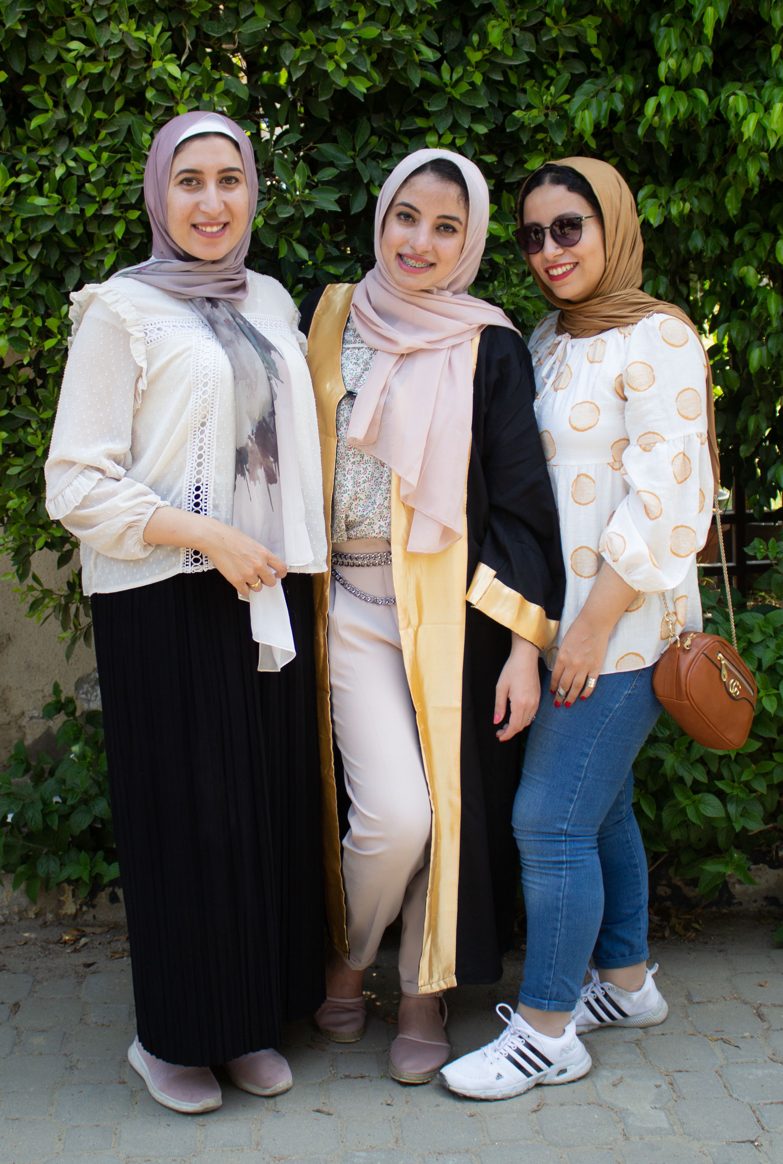 Three women in hijabs smiling together in casual clothing outdoors.