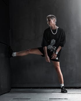 Full body androgynous female with short hair in black clothes with chain around neck standing in empty grey room and leaning foot against wall