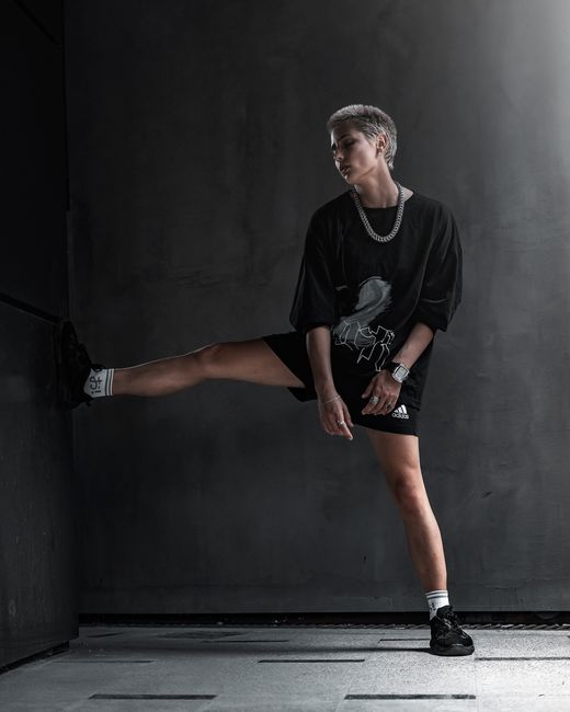 Full body androgynous female with short hair in black clothes with chain around neck standing in empty grey room and leaning foot against wall