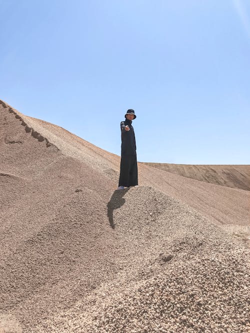 Unrecognizable male in long stylish outfit and hat standing and posing on dry sand in desert
