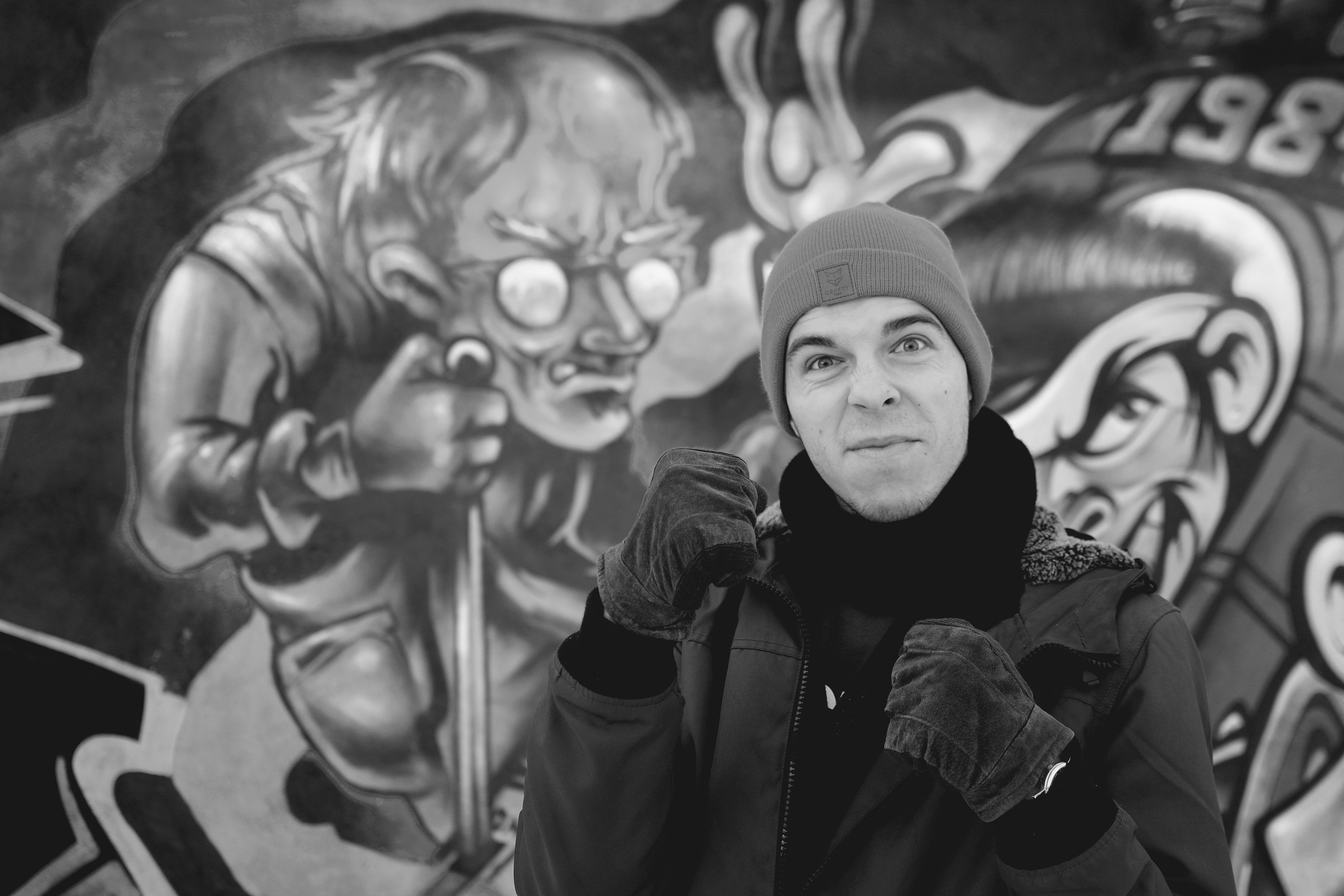 A man poses in a beanie and gloves against an urban graffiti backdrop.