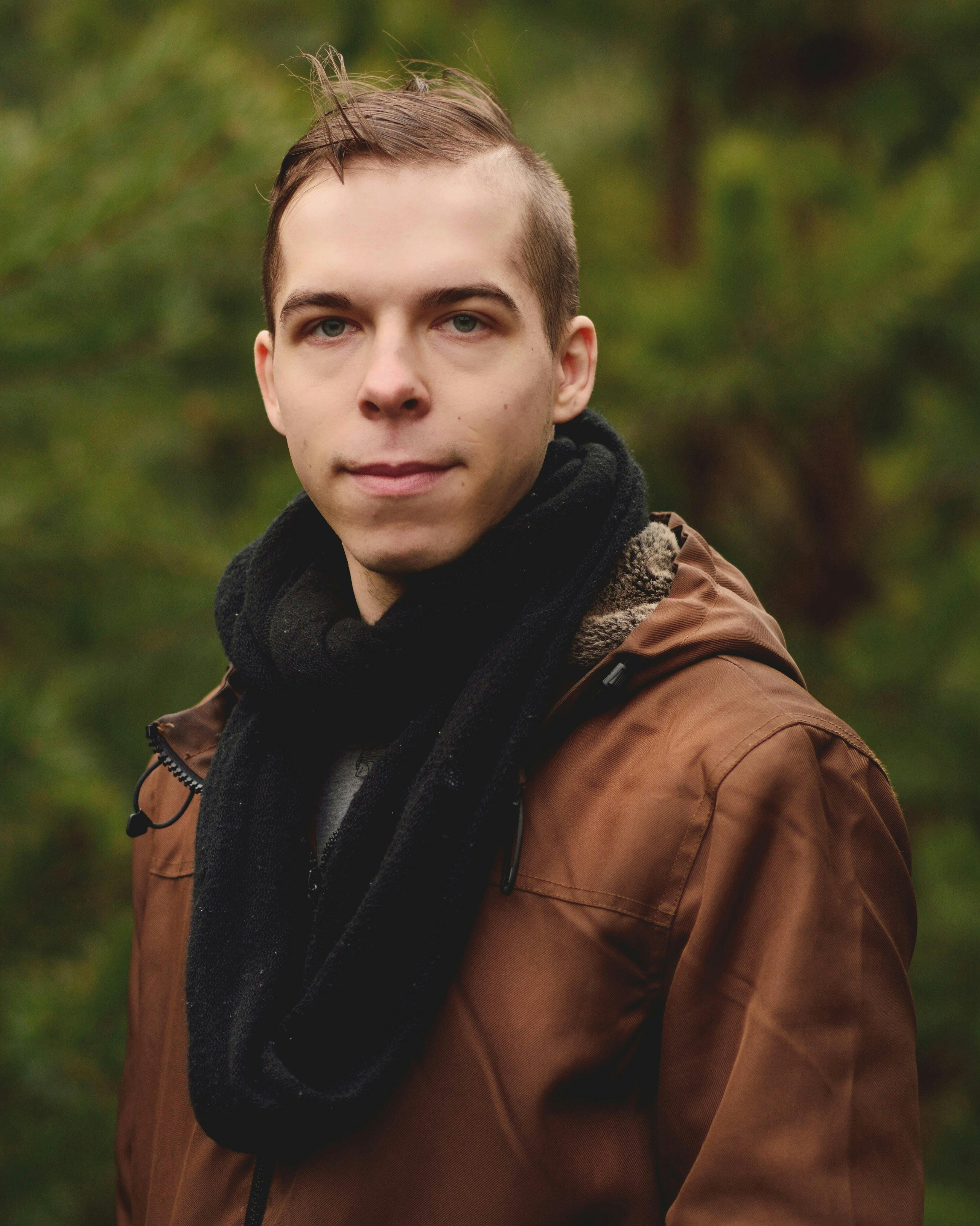 Young man outdoors wearing a brown jacket and black scarf in nature.