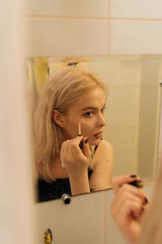 Woman applying makeup in front of bathroom mirror, focused expression, natural light.