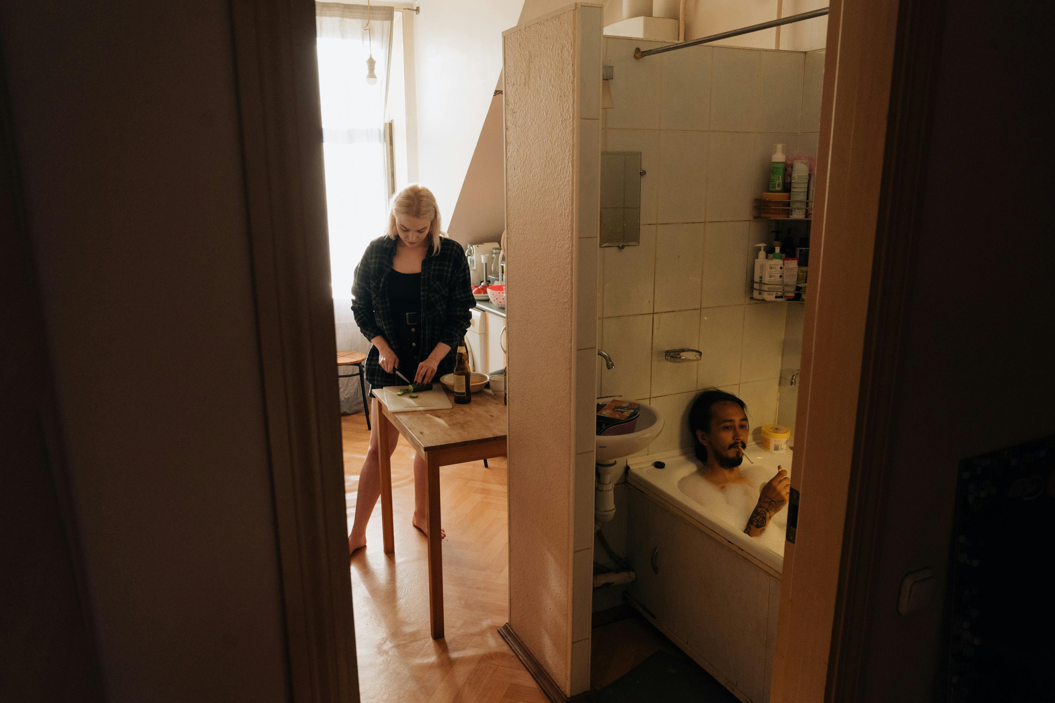 A woman prepares food in the kitchen while a man relaxes in a bathtub, capturing a cozy apartment lifestyle.