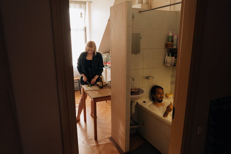 A Woman Preparing Food In The Kitchen While The Man Is Soaking Is Relaxing On The Bath Tub