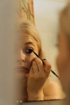Blonde woman applying eyeliner in front of a mirror, focused and precise.