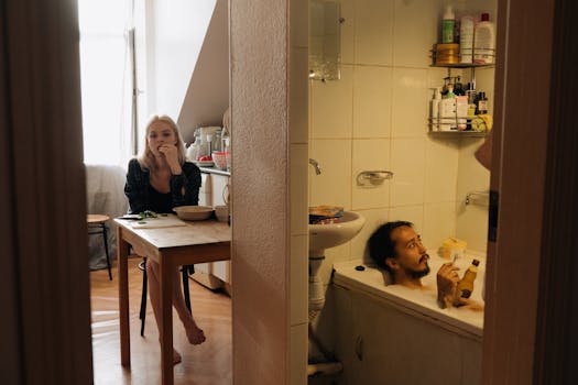 An intimate domestic moment shows a couple in a cozy apartment, one in the kitchen, another in the bathtub.