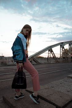 Fashionable woman poses in trendy outfit on a city bridge at sunset.
