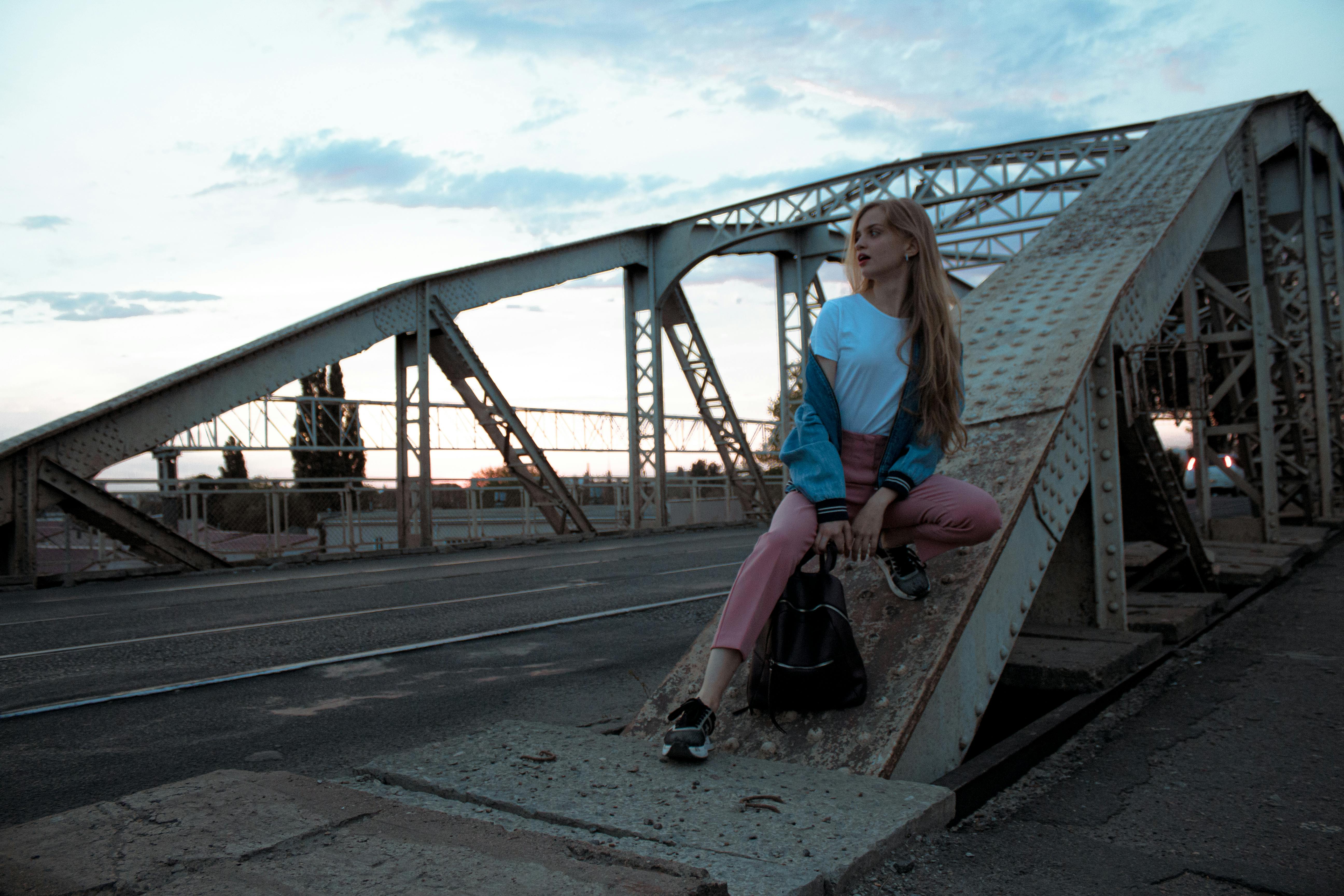 Stylish woman in casual outfit sits on a bridge during sunset, showcasing modern fashion.