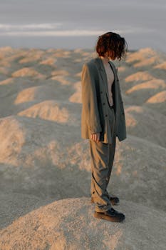 A model in a suit stands uniquely amidst a sandy desert under dawn light, capturing a minimalistic fashion statement.