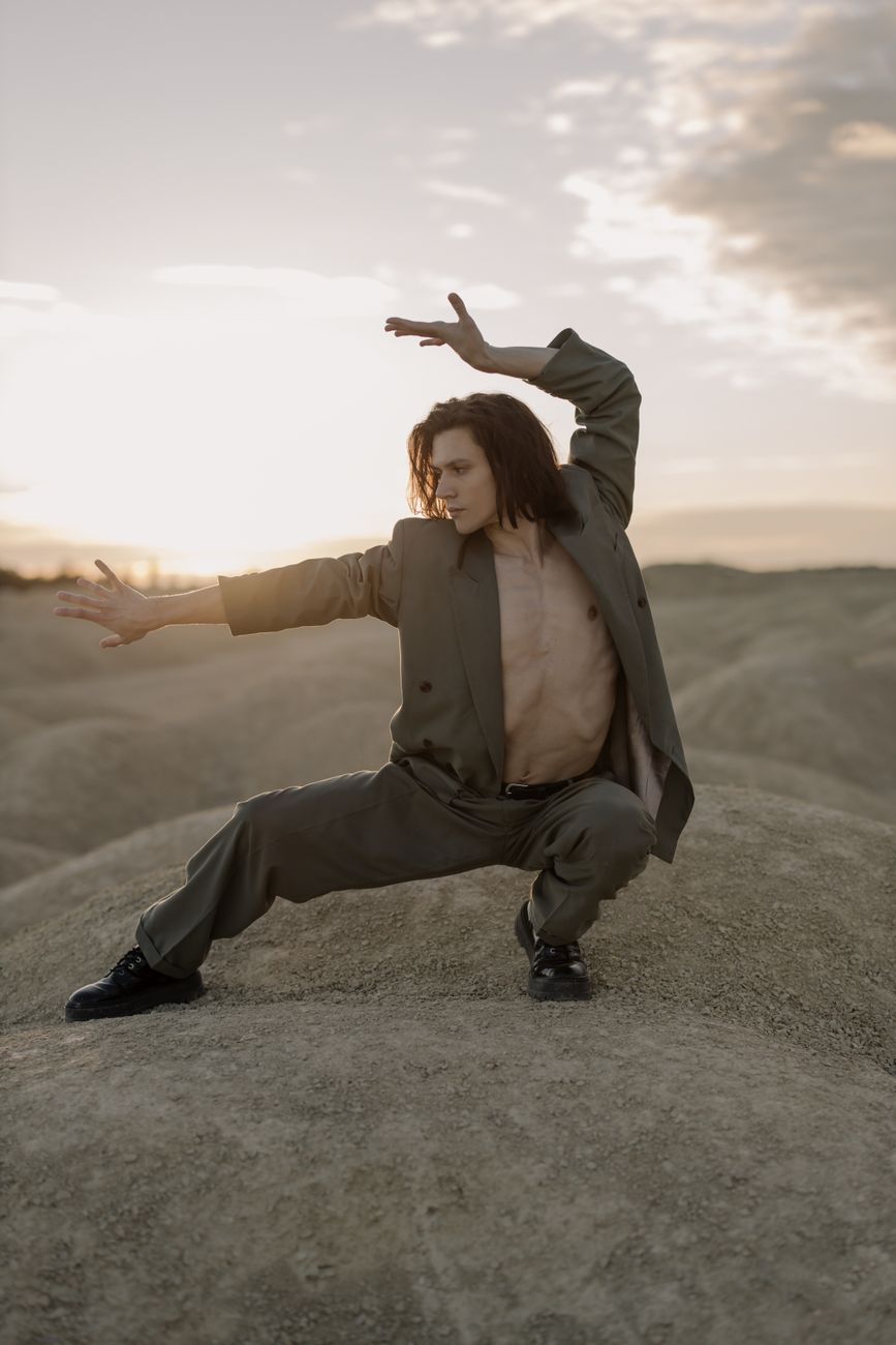 A shirtless dancer poses in a stylish suit on sandy dunes at sunset.