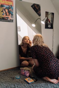 A young woman applies makeup while kneeling in front of a bedroom mirror.
