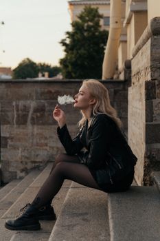 一名年轻女子穿着黑色皮夹克，坐在楼梯上，侧身吐着烟