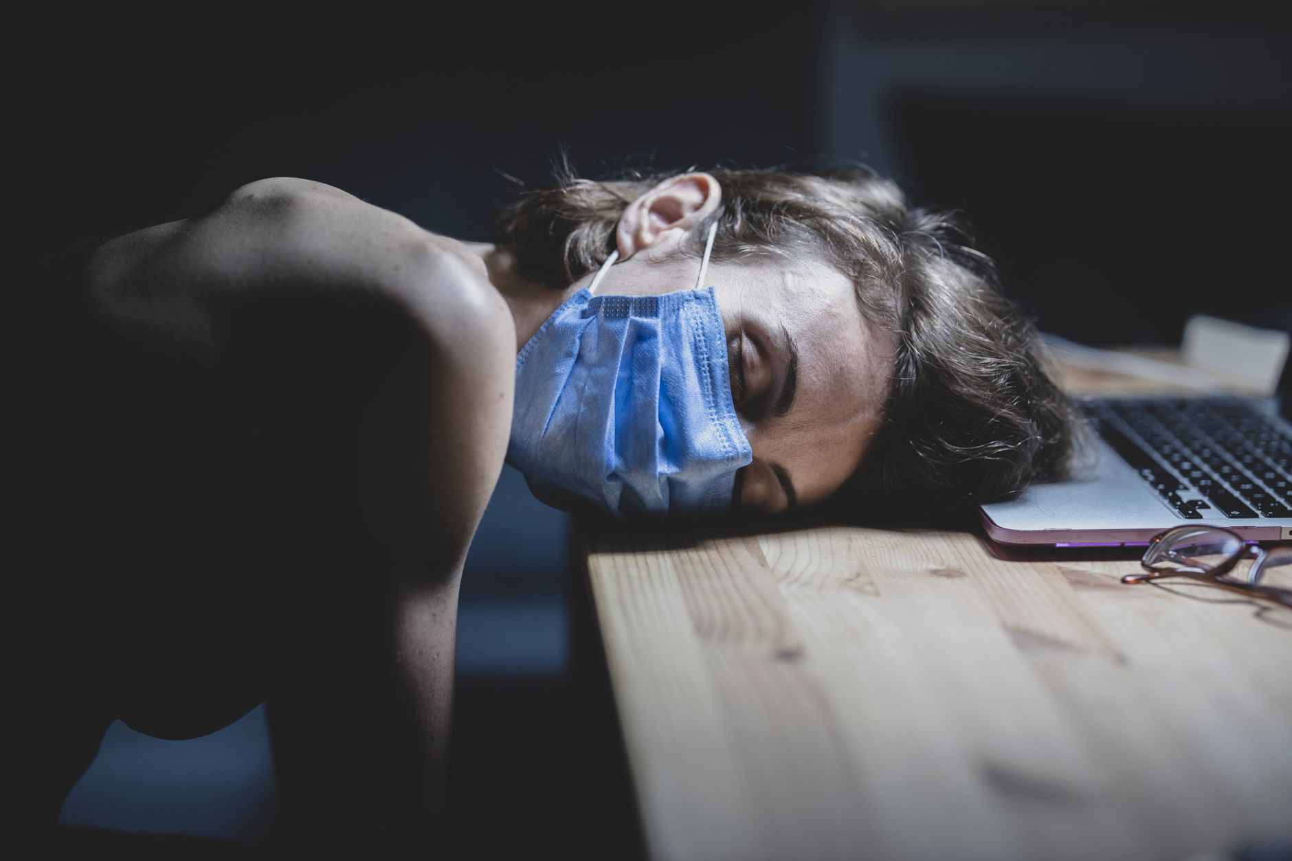 A woman in a mask sleeps on a desk, illustrating pandemic fatigue.