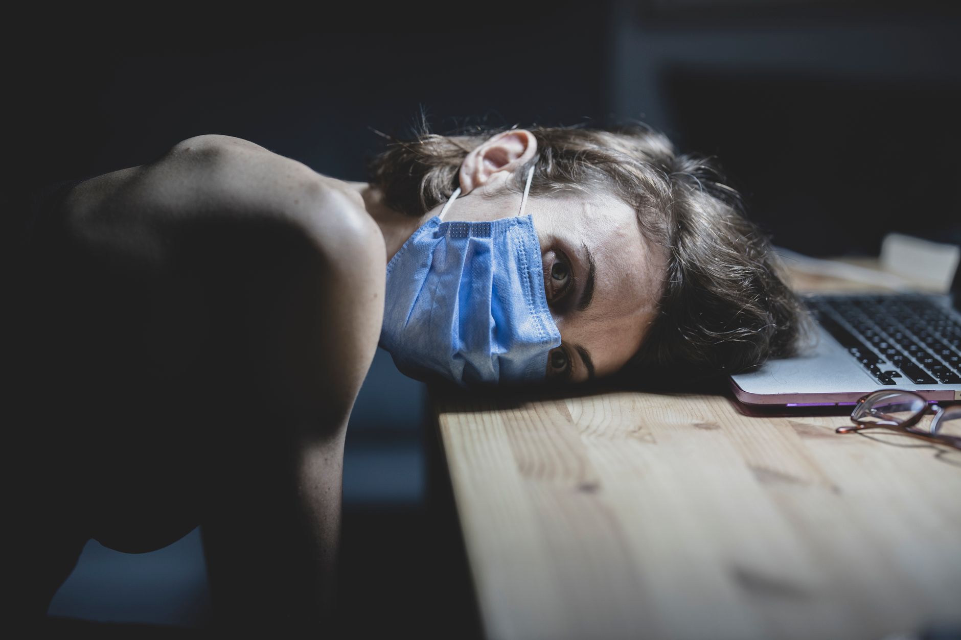 Woman wearing a face mask resting her head on a table beside a laptop, conveying pandemic fatigue and protection.