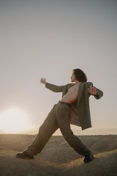 日落时分，一名身着西装的裸衣男子在沙丘上摆出动态姿势，将时尚与自然融为一体