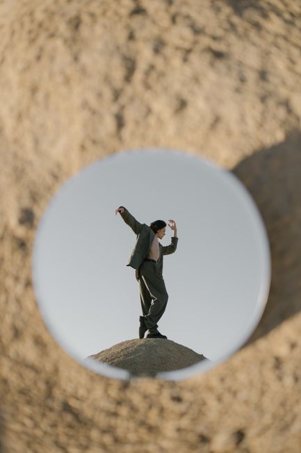 A dancer poses on a sandy hill, reflected in a round mirror in a desert setting.