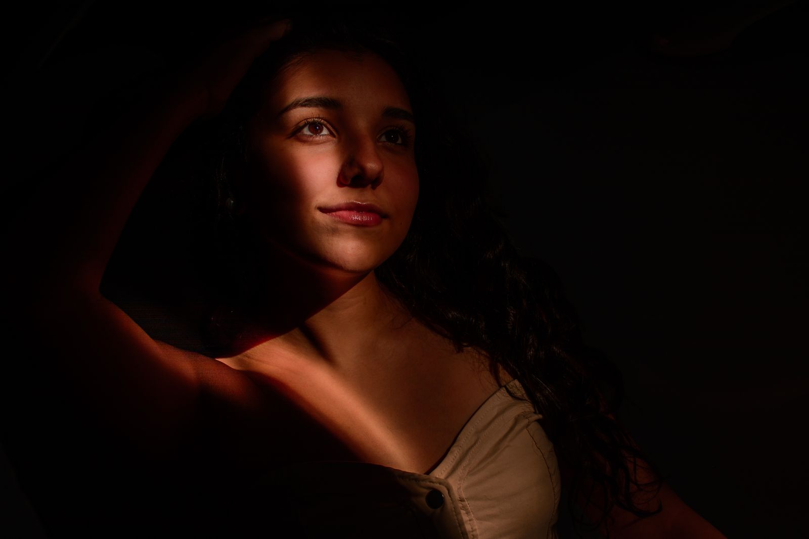 Woman with long dark hair in warm side light, arm raised, gazing upward against a dark background